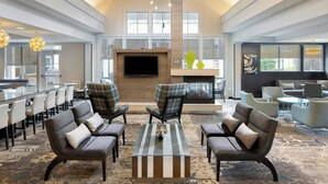 Eine geräumige Lobby mit hoher Decke, Kamin und Fernseher. Es gibt mehrere Sitzbereiche mit modernen Stühlen und einem zentralen Couchtisch.
