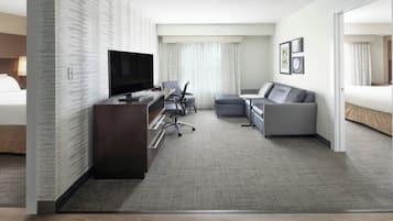 Una habitación de hotel moderna con una televisión de pantalla plana, un escritorio con silla, un sofá gris y una cama.