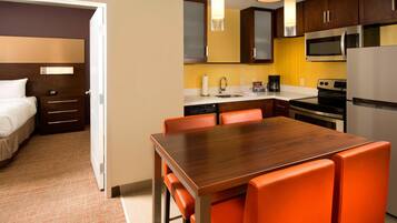 Habitación de hotel con una mesa de comedor de madera, sillas anaranjadas, una cama, una mesita de noche, una lámpara, un microondas, un refrigerador y una cafetera.