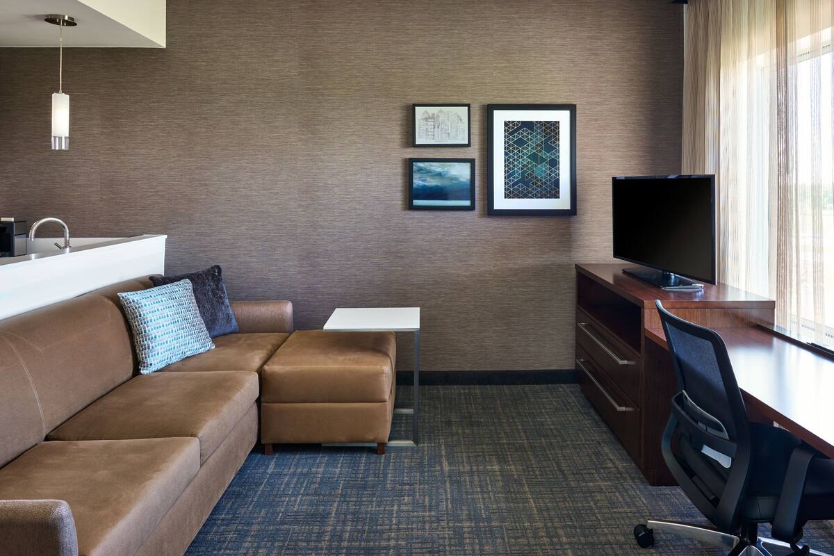 A hotel room with a brown leather sofa, a small white table, a TV, and a desk with a chair. There are framed pictures on the wall and a window with curtains.