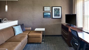 Habitación de hotel con sofá de cuero marrón, una mesita blanca pequeña, televisor y escritorio con silla. En la pared hay cuadros enmarcados y una ventana con cortinas.