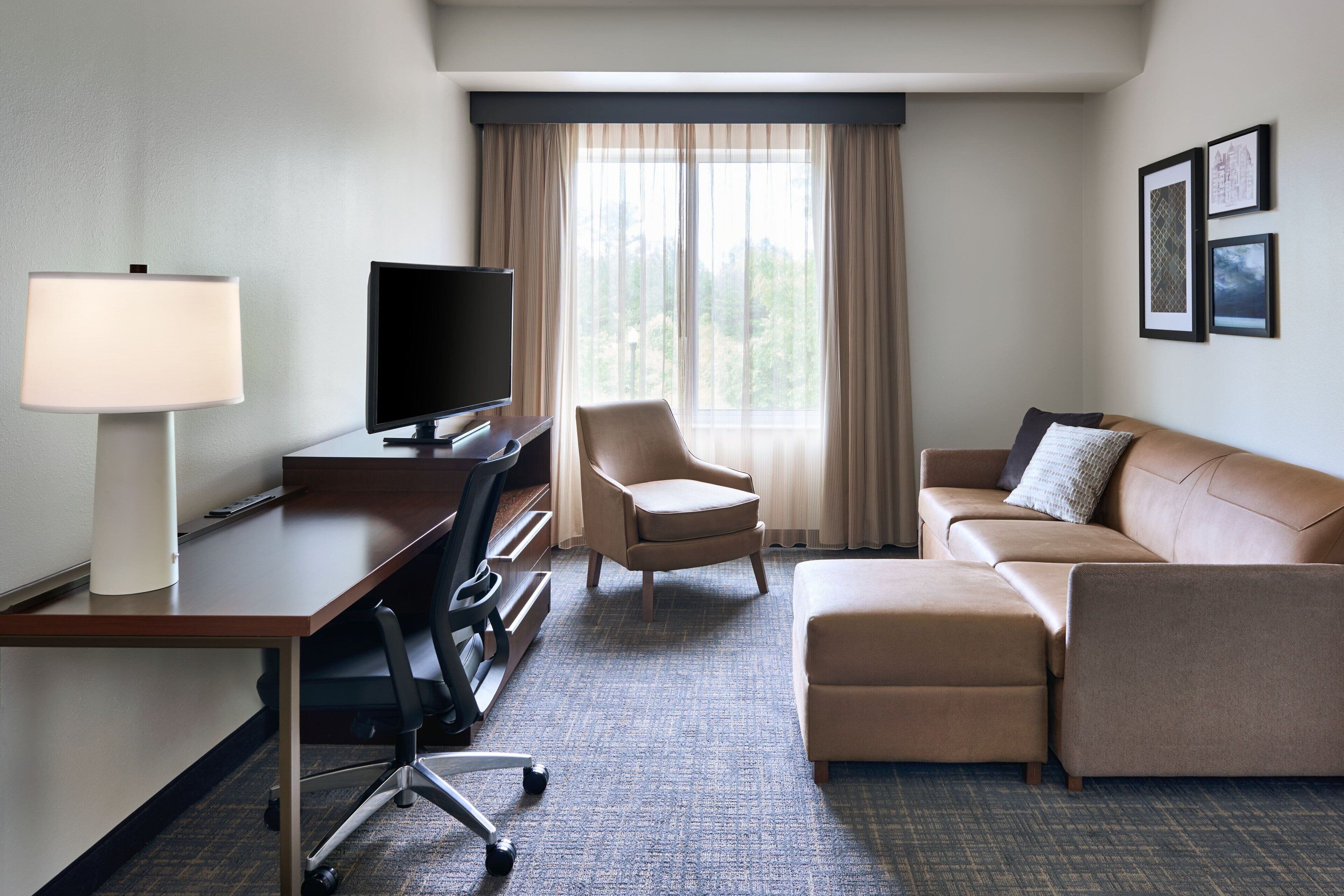 A hotel room with a desk, chair, television, sofa, and ottoman.