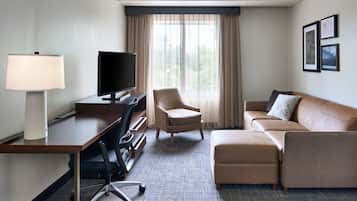 A hotel room with a desk, chair, television, sofa, and ottoman.