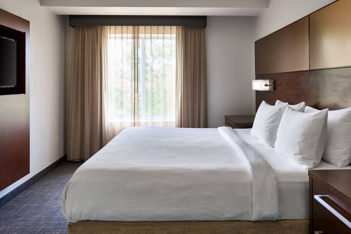 A hotel room with a large bed, a flat-screen TV mounted on the wall, a bedside table, and a window with curtains.