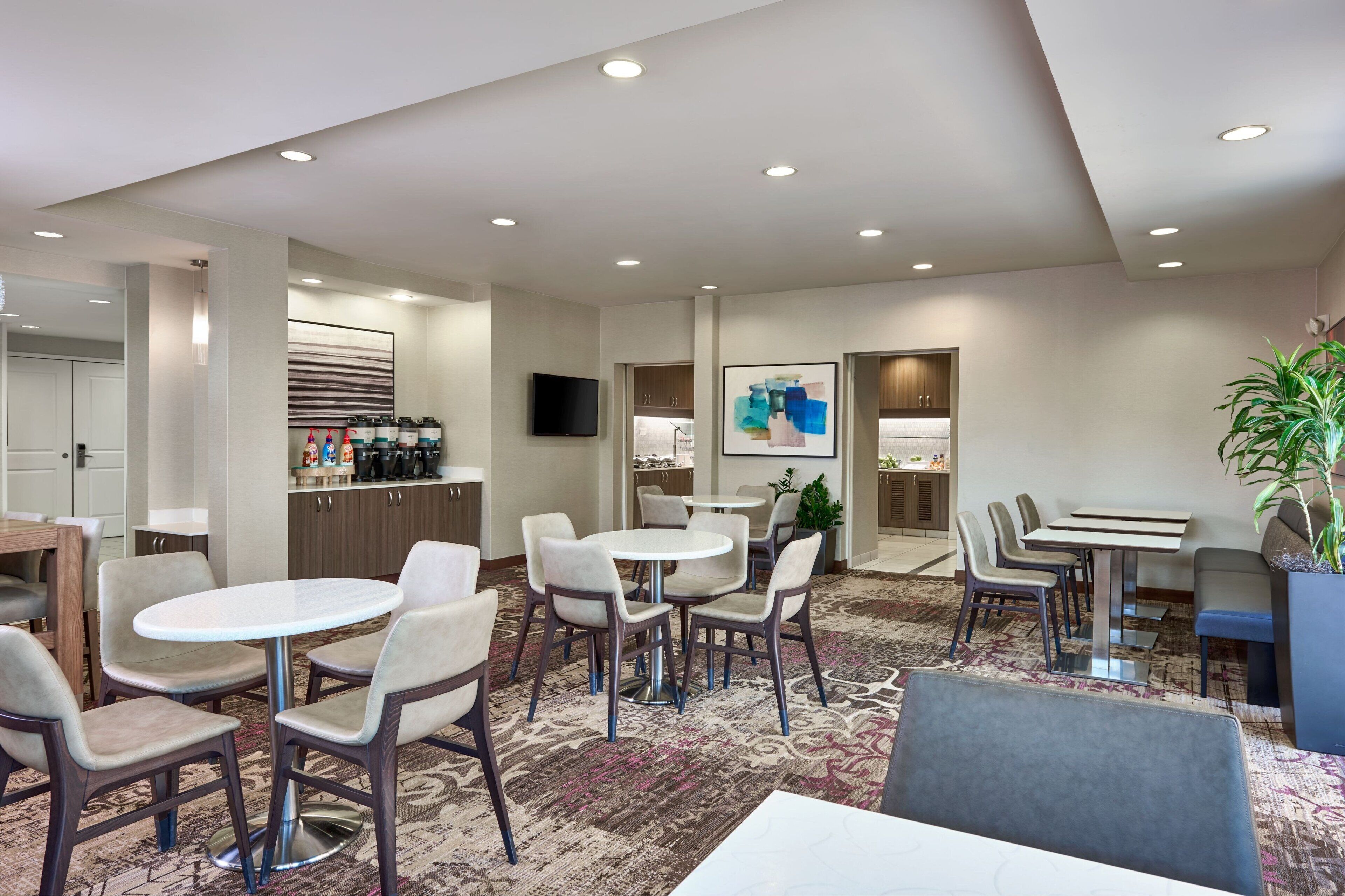 Een hotel lobby met ronde tafels en stoelen, een aan de muur bevestigde televisie en een koffiehoek.