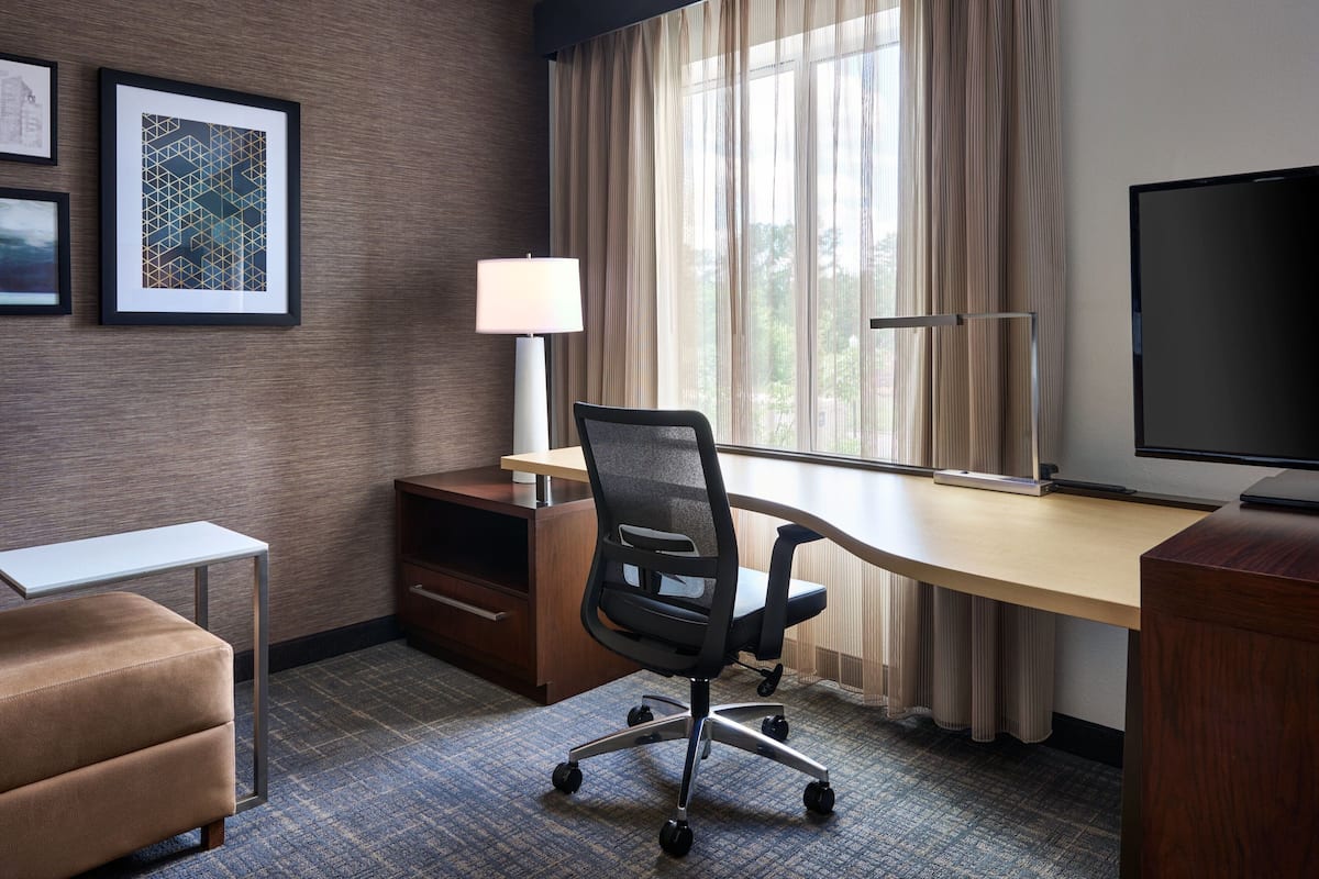 A hotel room with a desk, chair, lamp, TV, and a sofa.