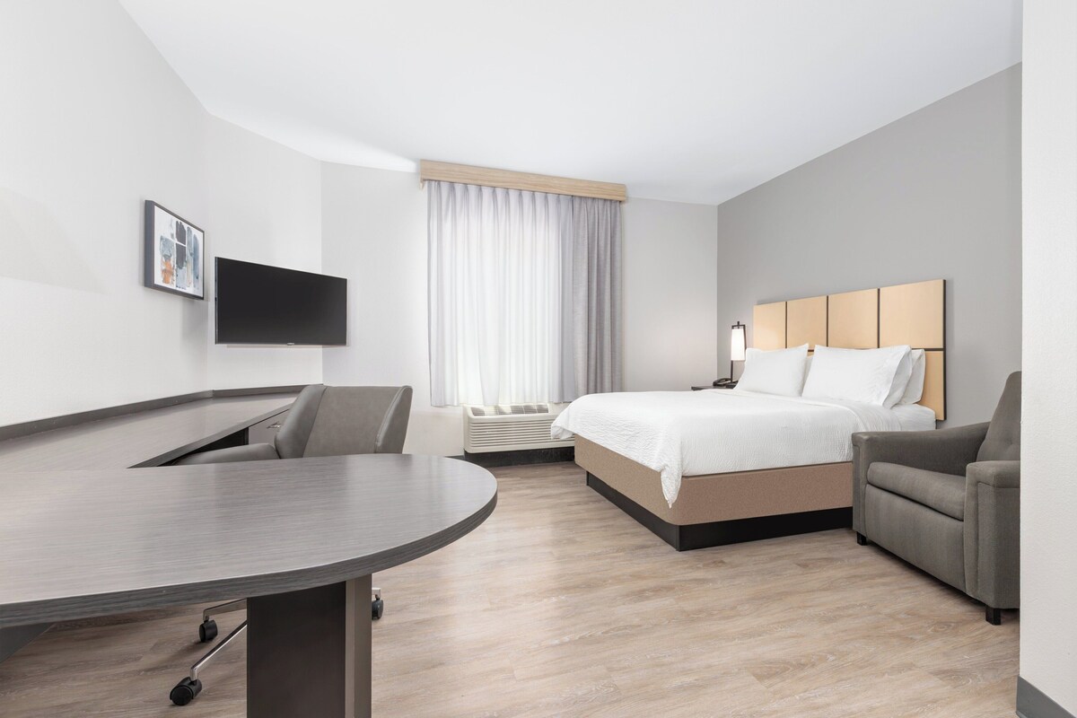 A modern hotel room with a bed, a desk, a chair, a flat-screen TV, and a large window with curtains.
