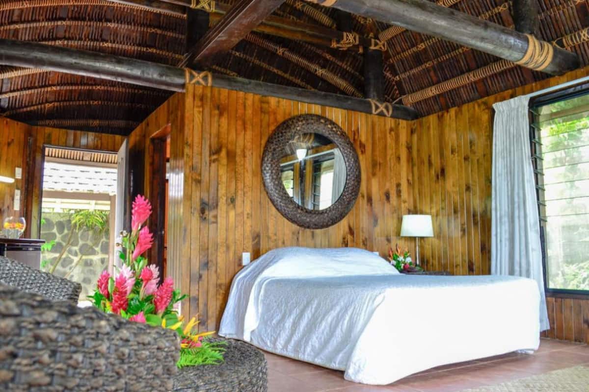 A wooden room with a bed, a round mirror, and a window.