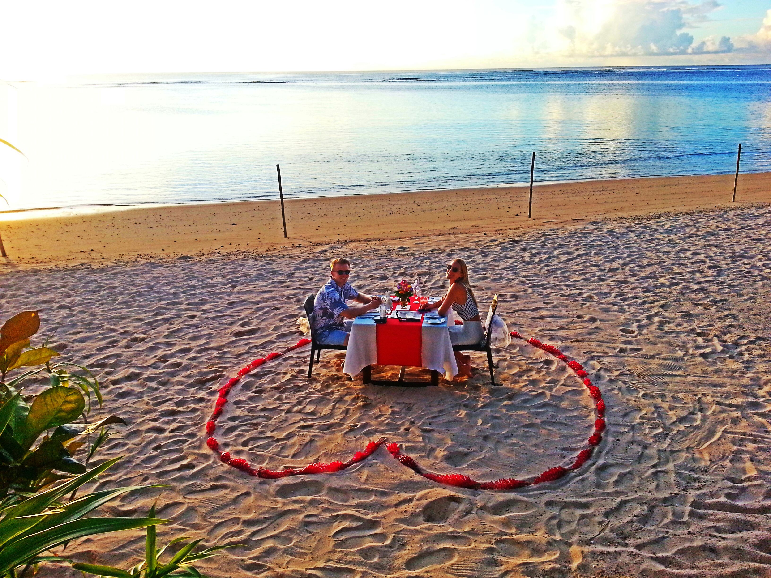 A couple dining at a table on a sandy beach with a view of the ocean.