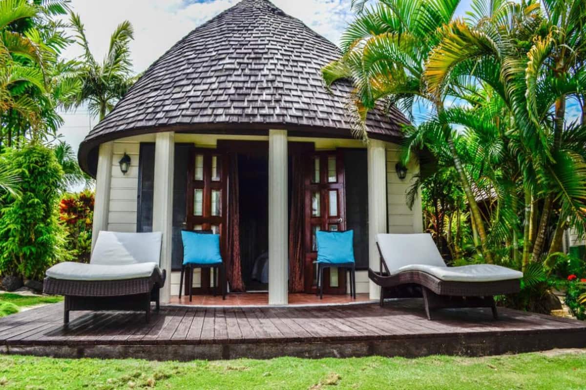 A thatched-roof structure with a porch, two lounge chairs, and a small table.