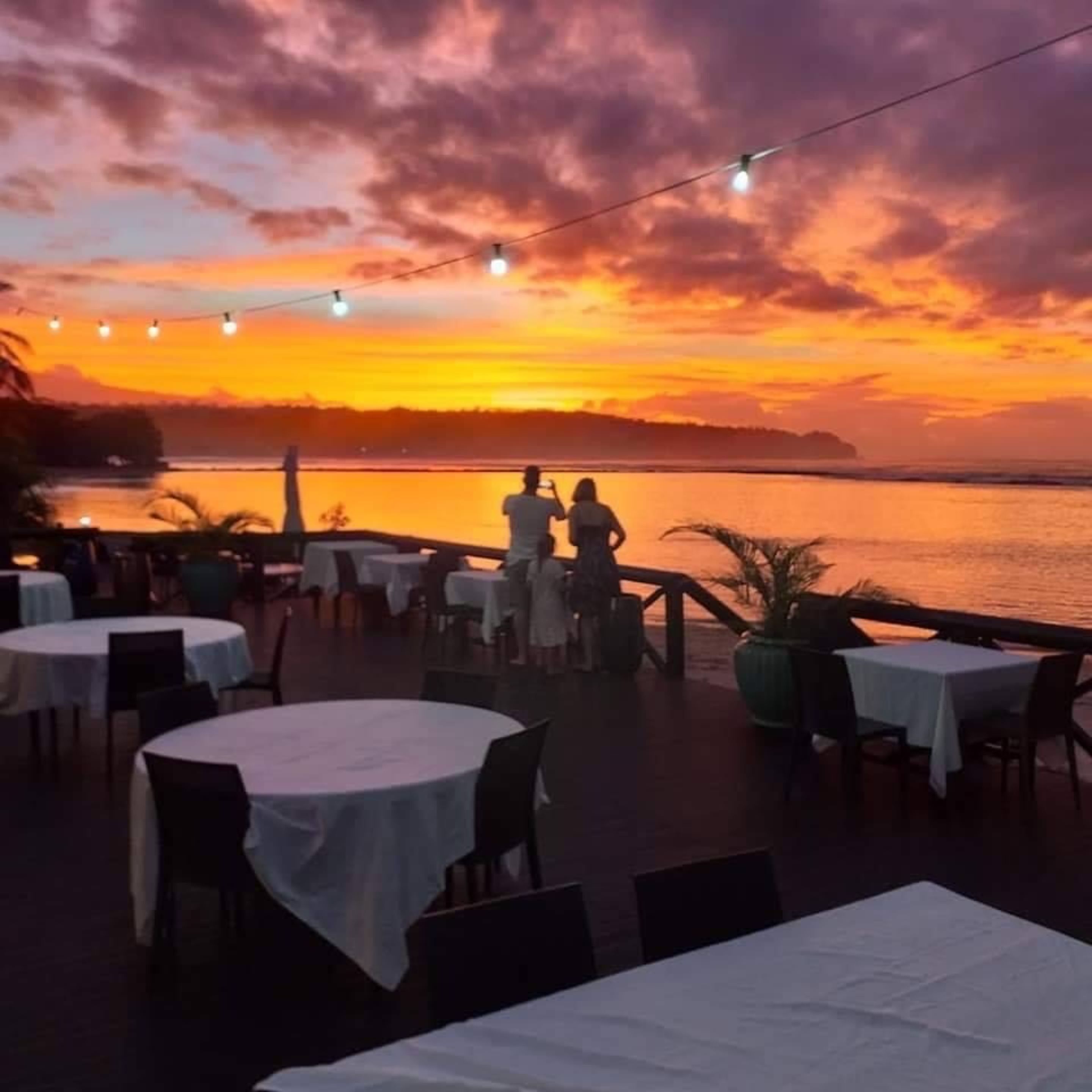 An outdoor dining area with tables and chairs, overlooking a body of water during sunset.
