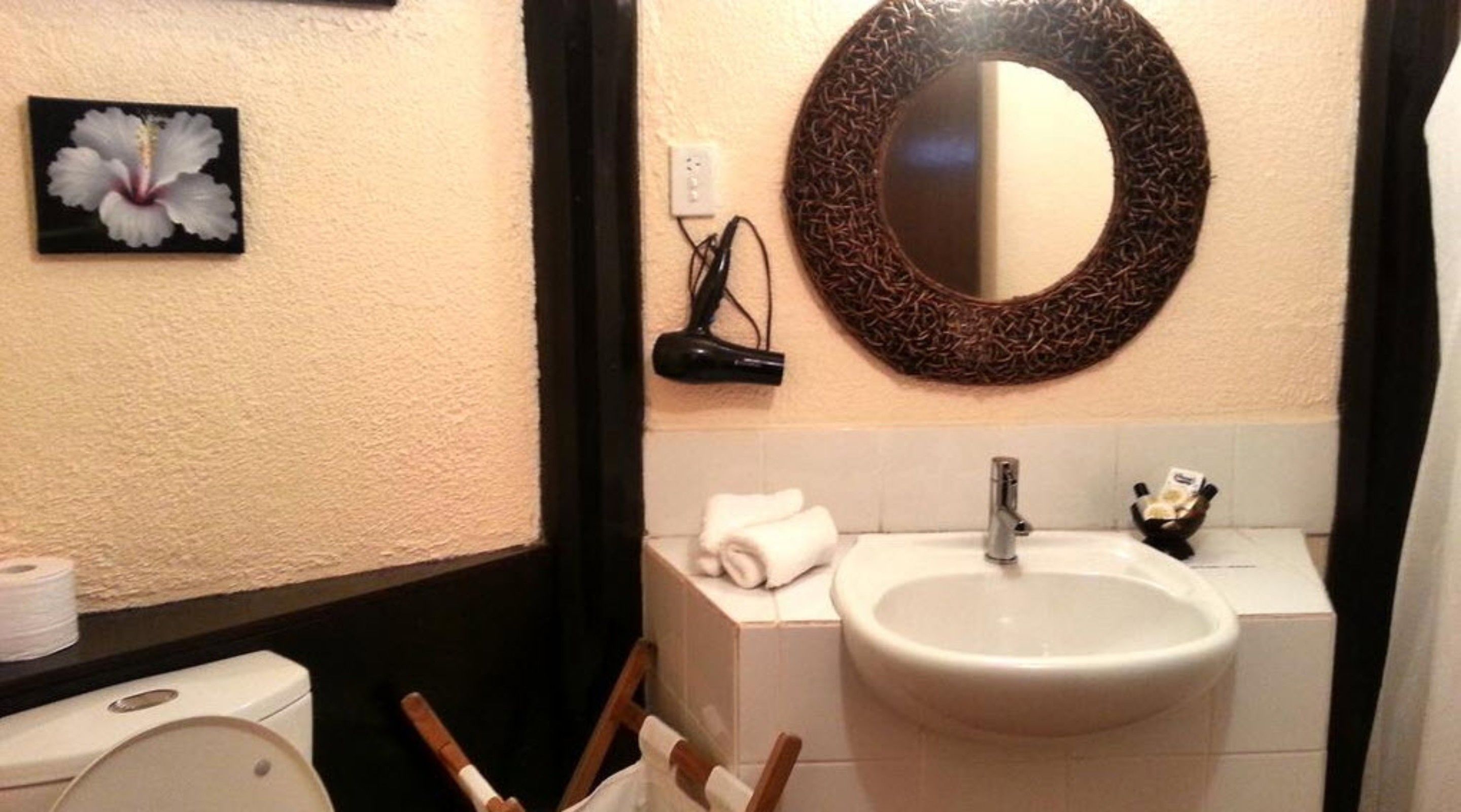 Ein Badezimmer mit Toilette, Waschbecken und einem Spiegel mit Korbflechtrahmen. Ein Fön ist an der Wand angebracht, und über der Toilette hängt ein Blumenbild.