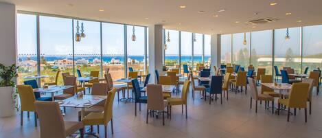 A spacious restaurant with large windows offering a view of the sea and buildings. Tables and chairs are neatly arranged.