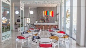 A modern bar area with a bar counter, colorful hanging lights, and a variety of seating options including round tables and chairs. - Nicholas Color Hotel (Ayia Napa)