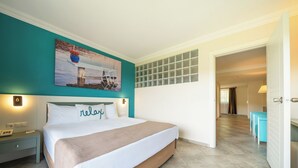 Ein Hotelzimmer mit Bett, Nachttisch, Wandbild, Fenster mit Jalousien und einer Tür zu einem weiteren Raum.