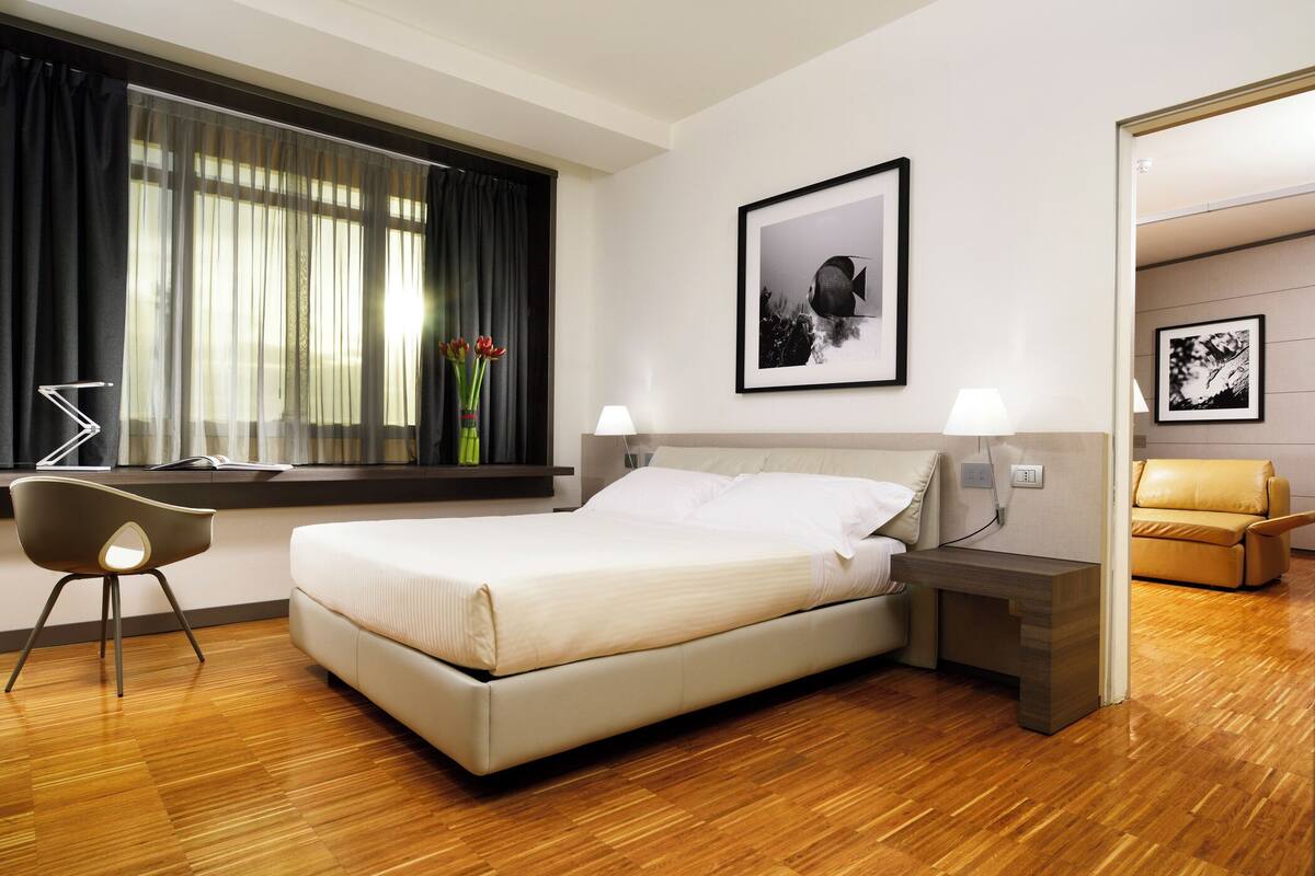 A modern hotel room with a large bed, bedside tables with lamps, a desk with a chair, and a window with sheer curtains.