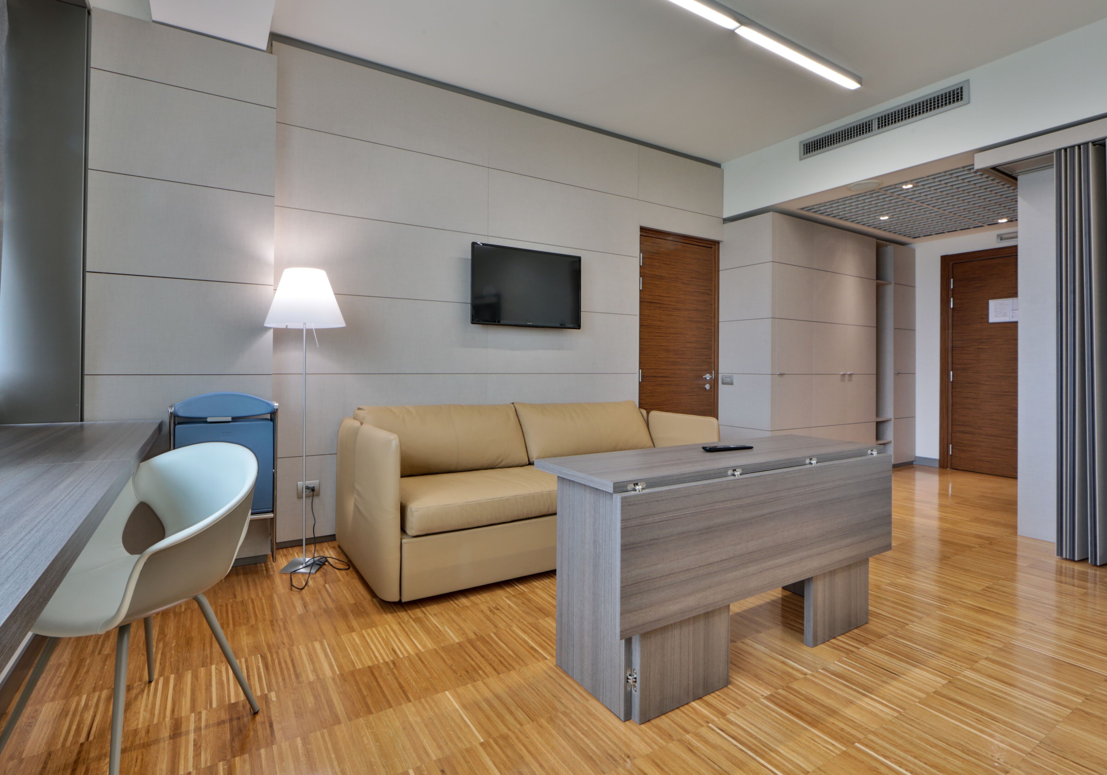 A modern office space with a desk, a sofa, a lamp, and a flat-screen TV mounted on the wall.