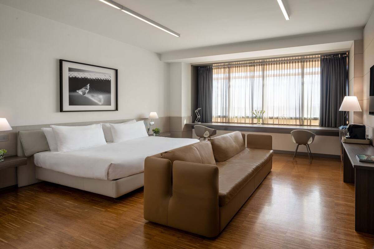 A modern hotel room with a large bed, a sofa, a desk, and a window with curtains.