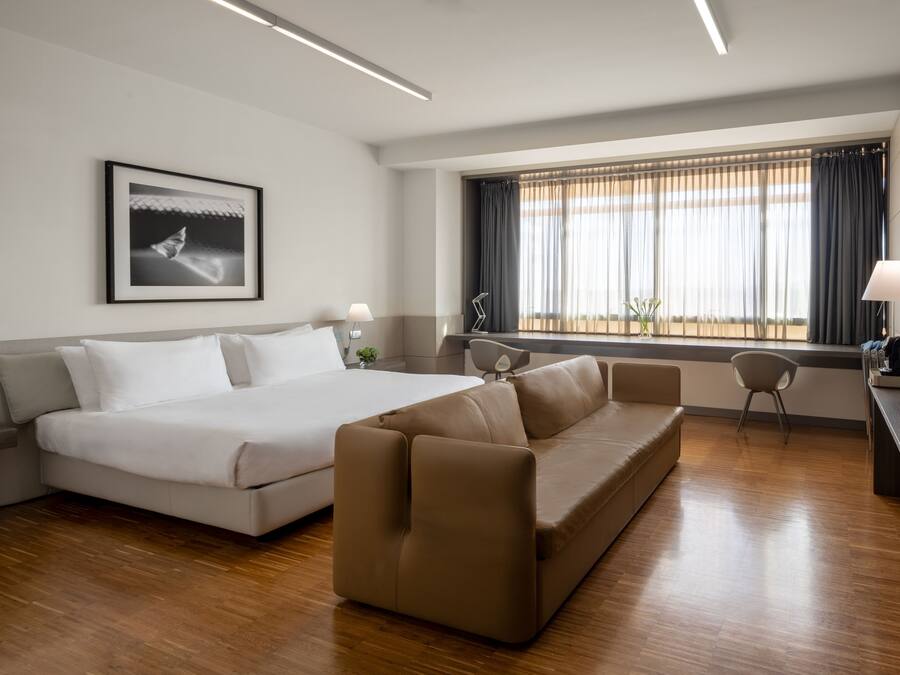 A modern hotel room with a large bed, a sofa, a desk, and a window with curtains.