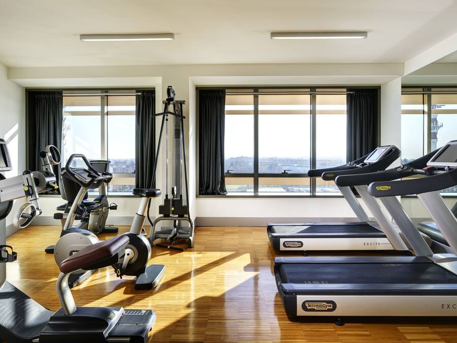 A modern gym with cardio equipment, mirrors, and large windows offering a city view.