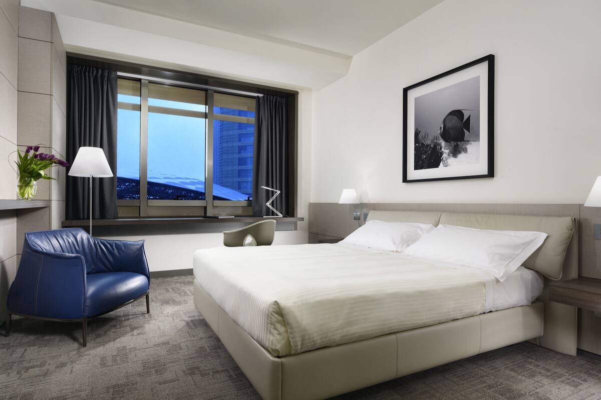 A hotel room with a large bed, a desk, a chair, a window with curtains, and a framed picture on the wall.