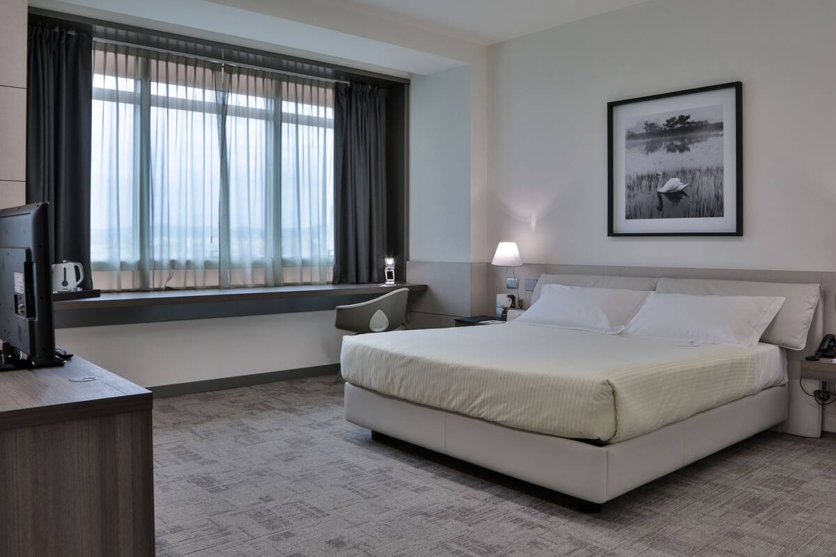 A hotel room with a bed, bedside lamps, a desk with a computer, a window with curtains, and a framed picture on the wall.