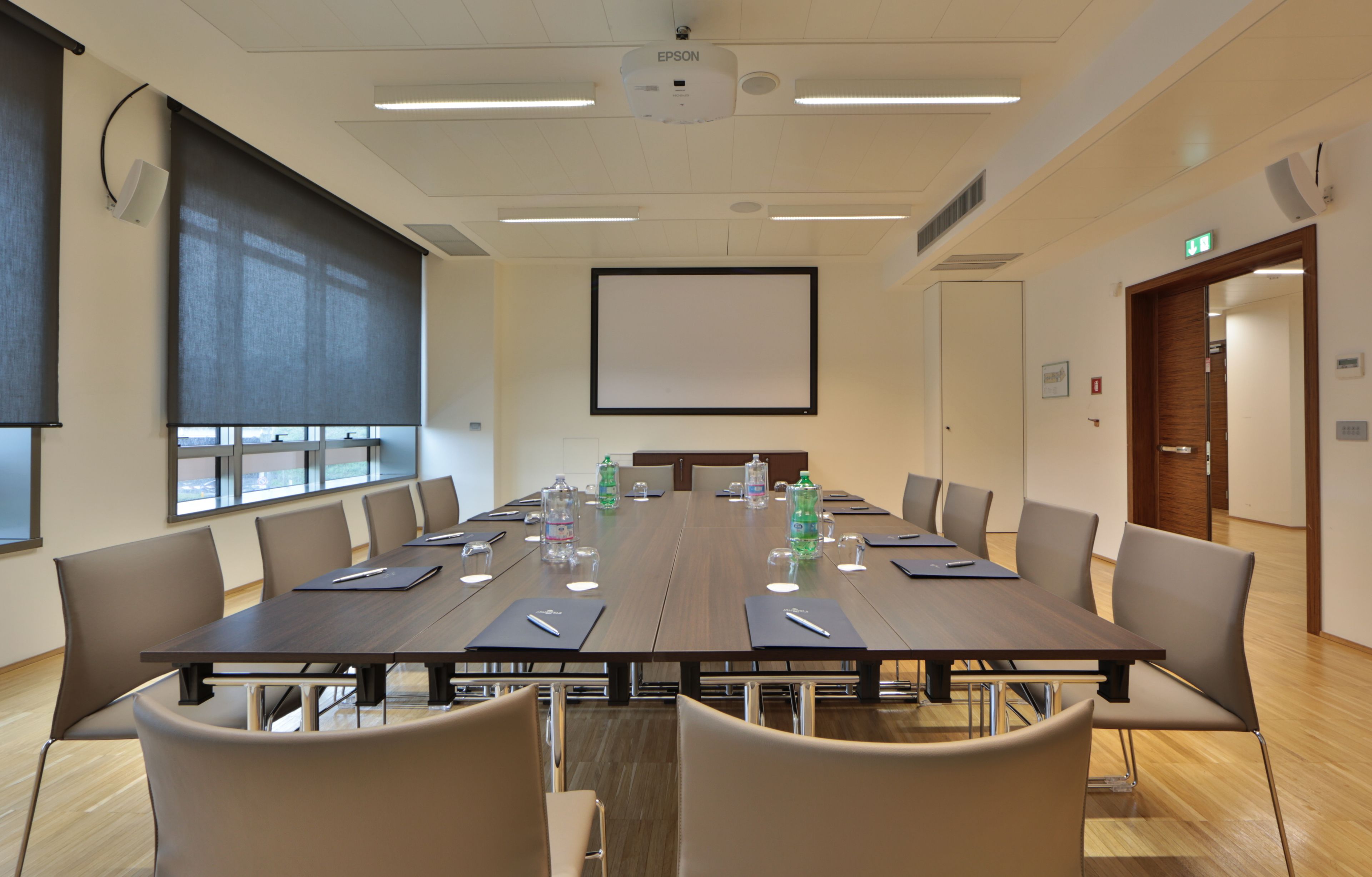 A conference room with a long table, chairs, water bottles, and a projector screen.
