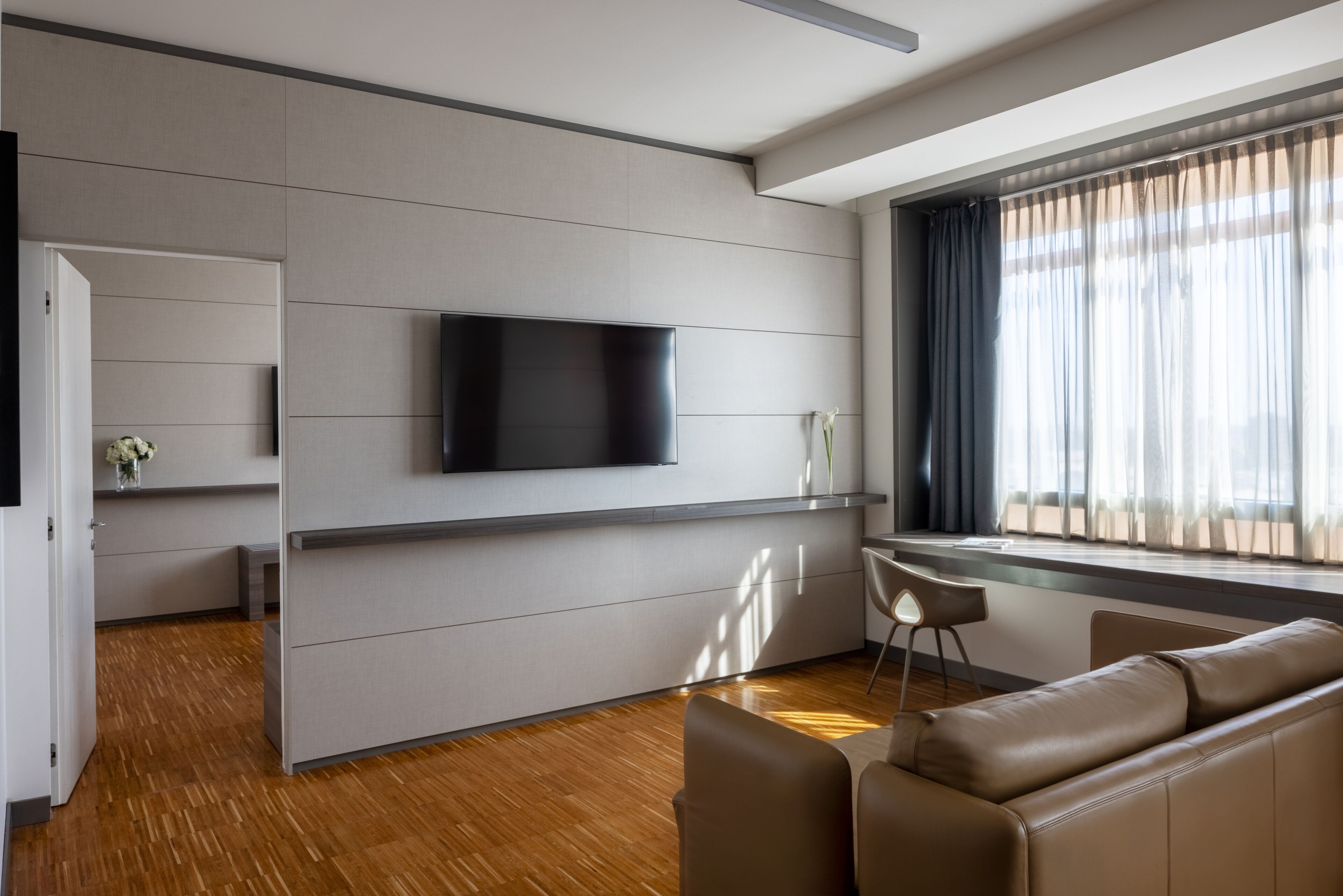 Una habitación de hotel moderna con un televisor de pantalla plana montado en la pared, un rincón de descanso con sofá y sillones, piso de madera y una ventana con cortinas.