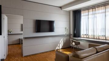 Una habitación de hotel moderna con un televisor de pantalla plana montado en la pared, un rincón de descanso con sofá y sillones, piso de madera y una ventana con cortinas.