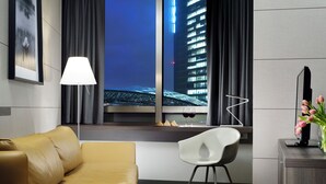 A modern hotel room with a leather sofa, a white chair, a desk with a vase of flowers, a lamp, and a view of a cityscape at night. - UNA Hotels San Vitale Bologna (Bologna)