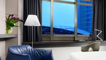 Quarto de hotel moderno com uma janela grande, uma poltrona de couro azul, uma mesinha de cabeceira com abajur e uma cama com cabeceira branca.