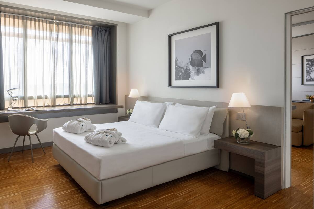 A modern hotel room with a large bed, two bedside tables with lamps, a chair, and a window with curtains.