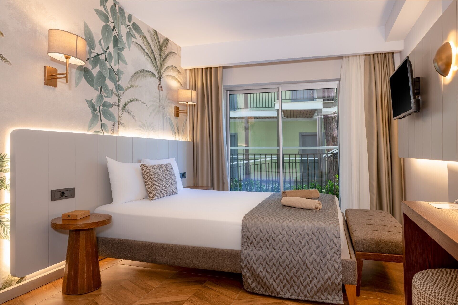 A modern hotel room with a large bed, a bedside table, a chair, a TV mounted on the wall, and a balcony with a view of the building.