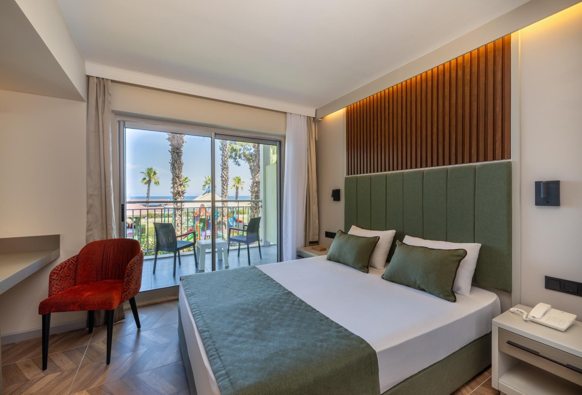 Ein Hotelzimmer mit einem großen Bett, einem Sessel, einem Nachttisch mit Telefon sowie Blick auf Palmen und einen Spielplatz.