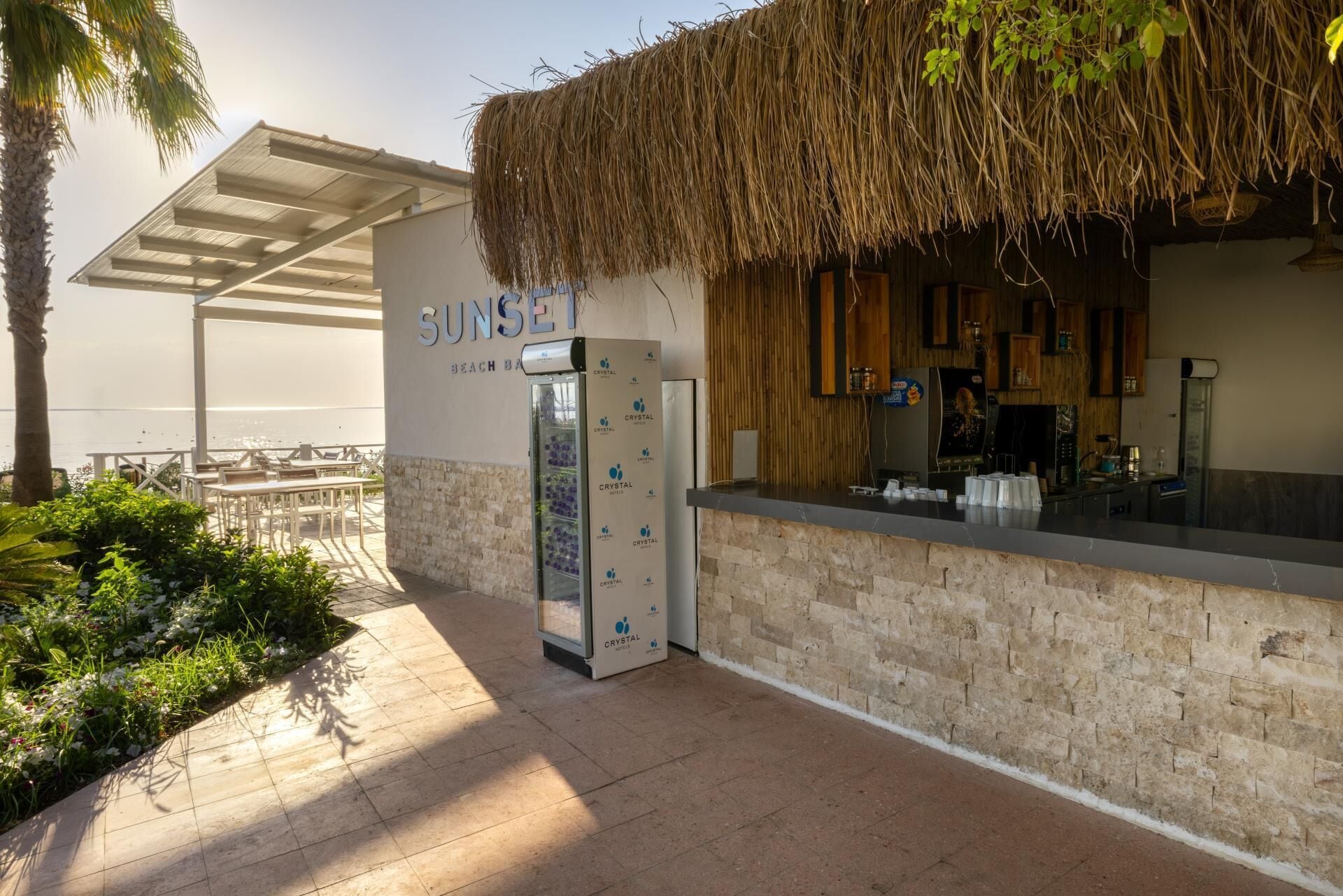 A beach bar with a thatched roof, a sign reading 'SUNSET BEACH BAR,' and a visible counter.