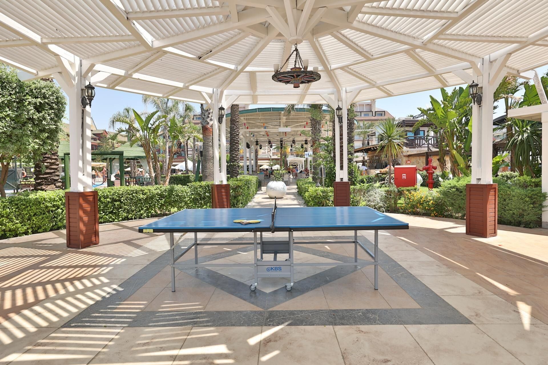 Un espacio al aire libre techado con mesa de ping pong, palmeras y una estructura tipo pabellón.