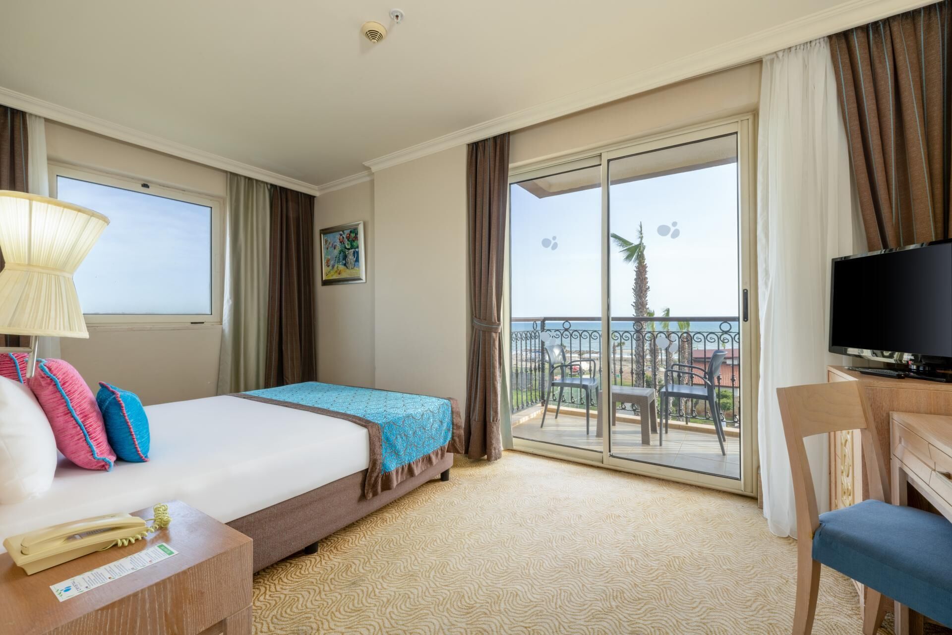 A hotel room with a bed, a desk with a telephone, a television, and a balcony with a view of the sea.