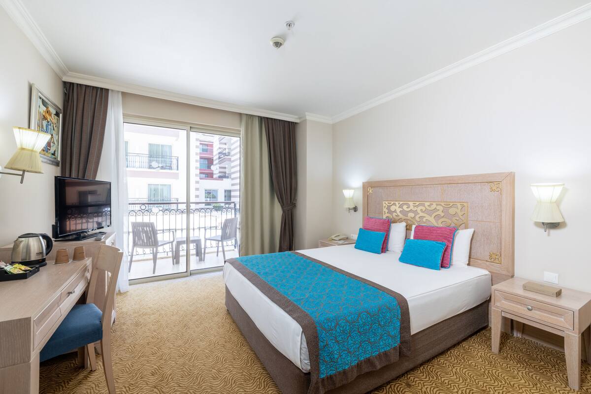 A hotel room with a bed, a desk with a chair, a television, a balcony with a view of buildings, and a window with curtains.