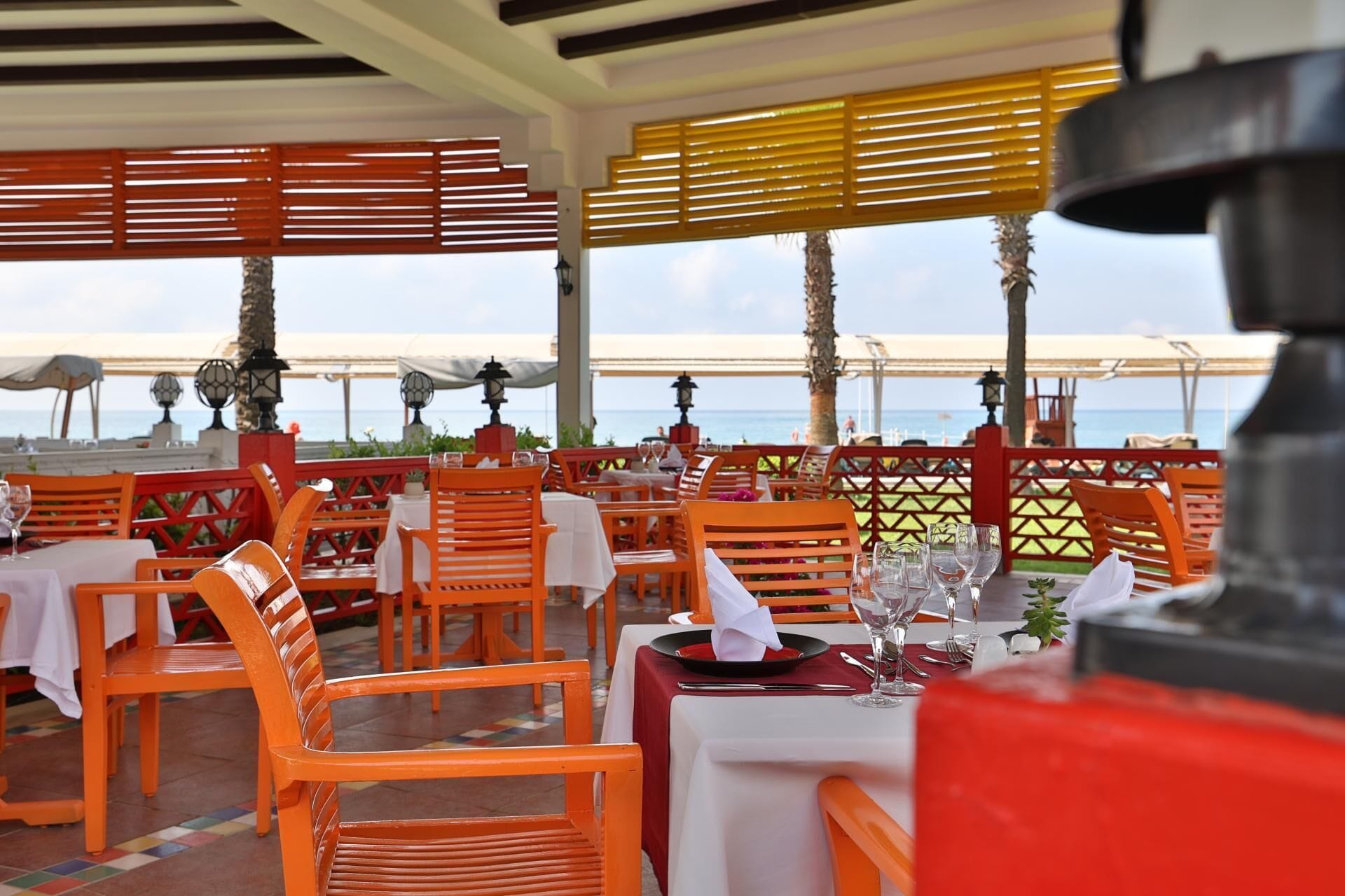 An outdoor restaurant with orange chairs and tables, a view of the sea, and a red and yellow striped awning.