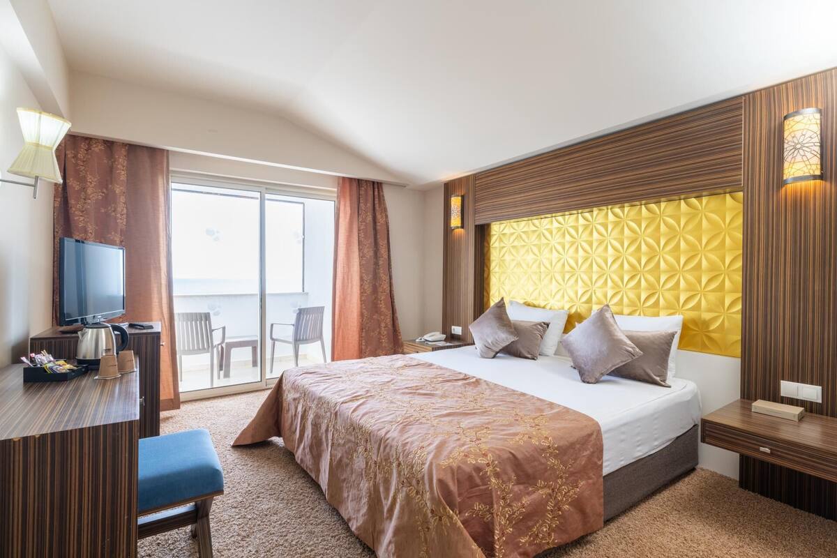 A hotel room with a bed, a desk with a kettle, a chair, a TV, and a balcony with curtains.