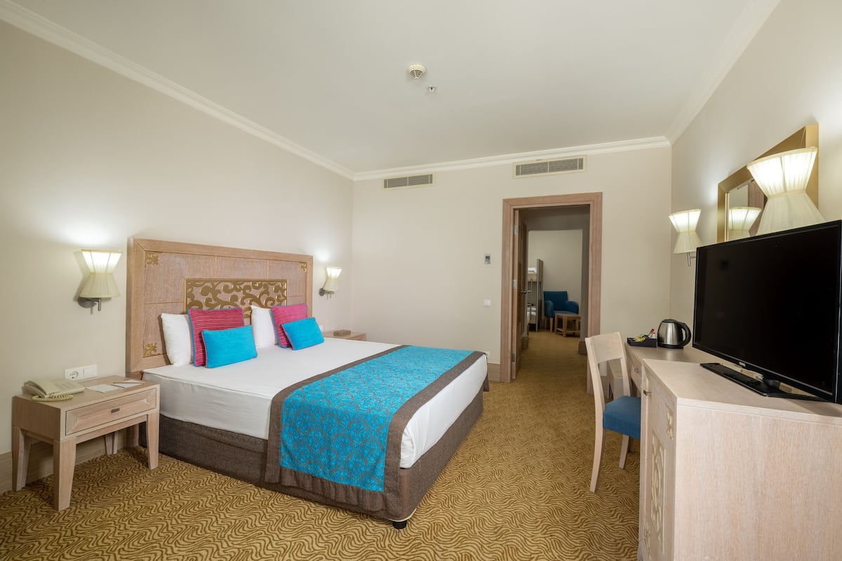 A hotel room with a bed, bedside table, lamp, TV, and a desk with a chair.