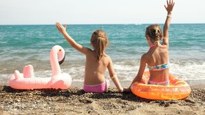 Zwei Kinder sitzen am Kiesstrand, eines mit einem rosa Schwimmflamingo und das andere mit einem orangenen Schwimmreifen, beide zum Meer zugewandt.