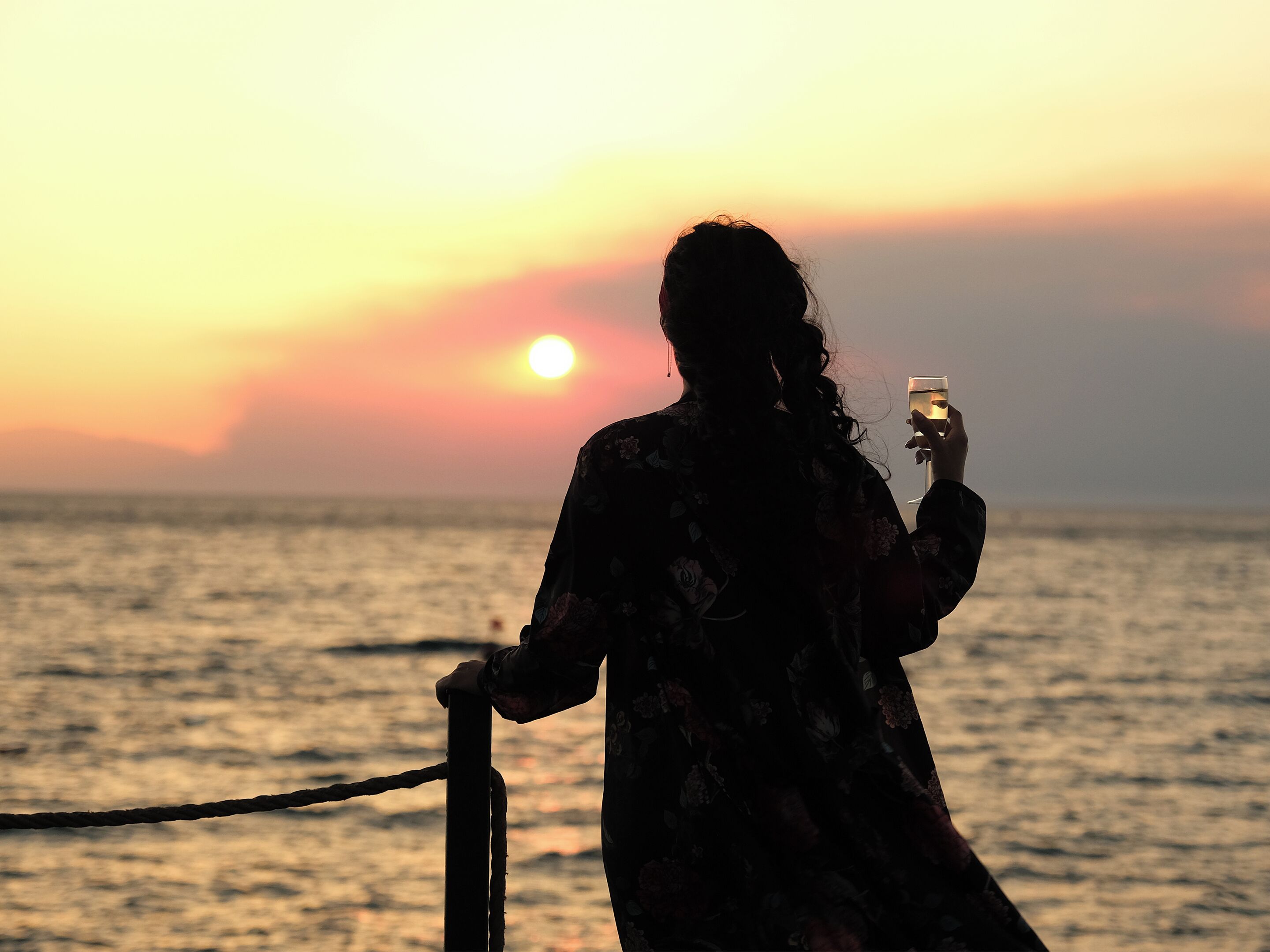 En person holder et glas og står ved et gelænder med solnedgang i baggrunden.