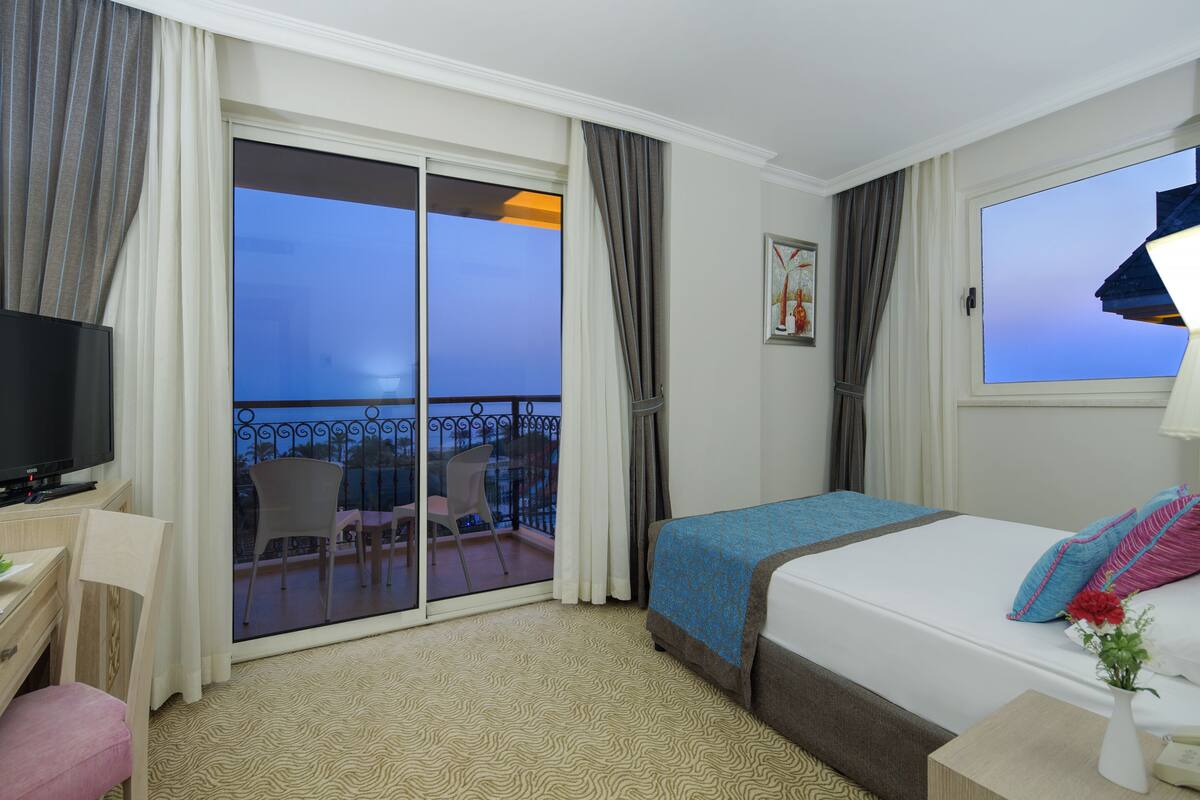 A hotel room with a bed, a desk with a TV, a balcony with chairs, and a view of the sky.
