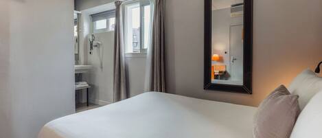 A hotel room with a bed, a mirror, and a view of a bathroom with a shower and a sink.