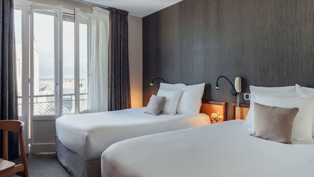 Habitación de hotel con dos camas, una silla de madera, una mesita de noche con lámpara y vistas por un ventanal grande.