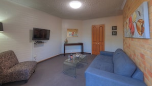 A living room with a TV, sofa, patterned armchair, coffee table, and artwork on the walls. - Cousins Motor Inn (Inverell)