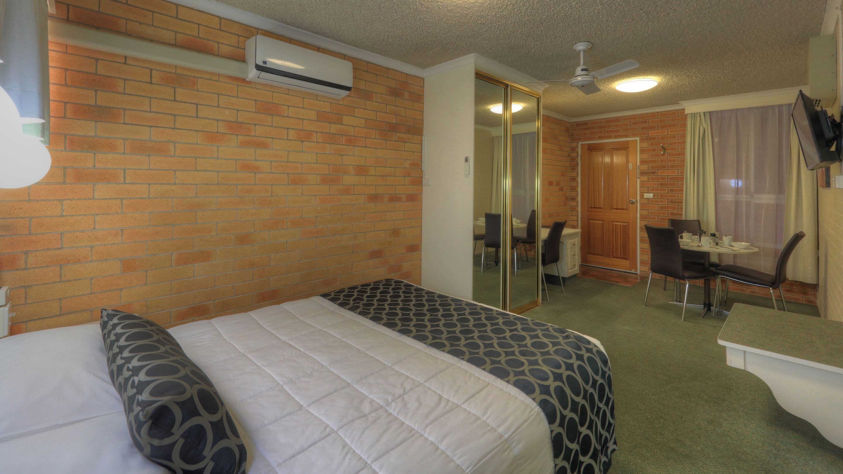A hotel room with a brick wall, a bed with patterned bedding, a desk with a chair, a mirror, and a ceiling fan.