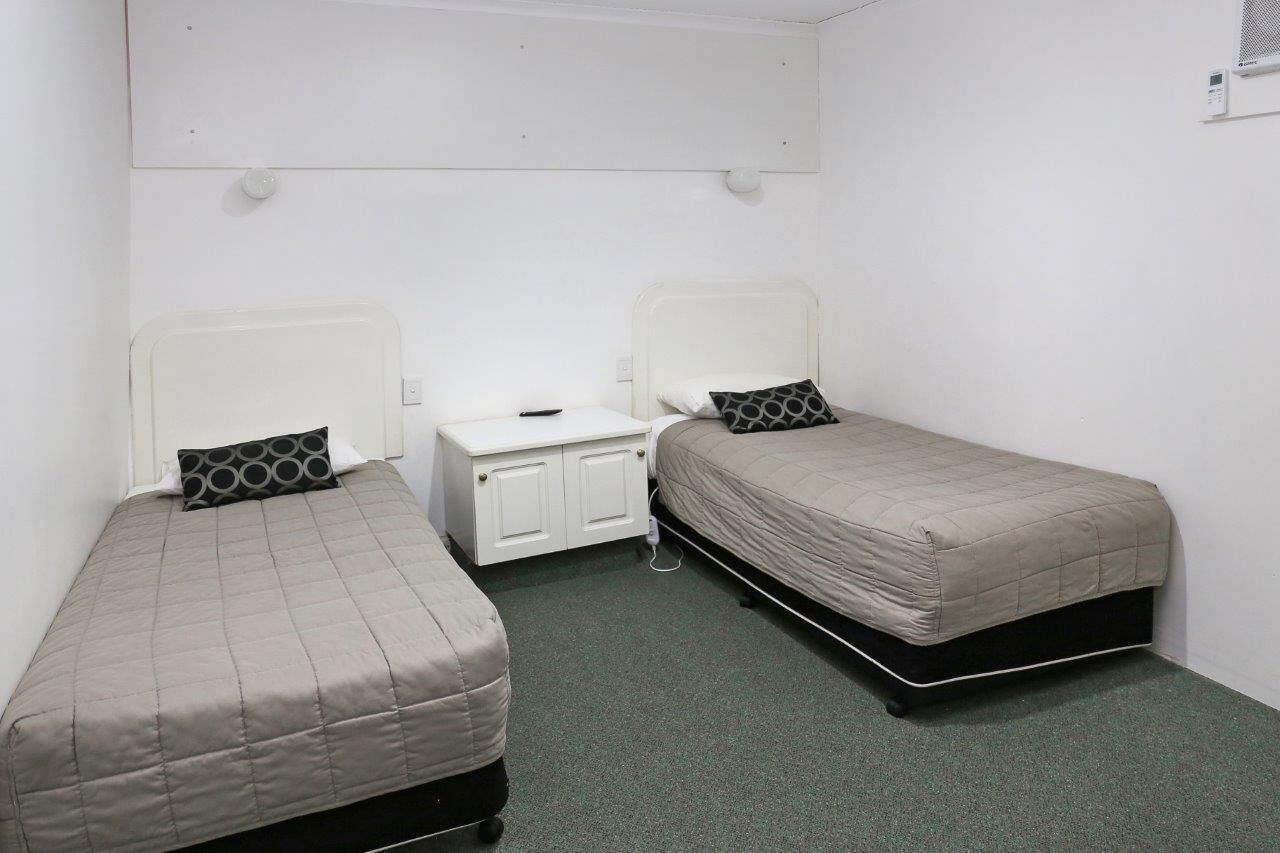 A small, single-room accommodation with two single beds, a small white bedside table, and a wall-mounted air conditioning unit.