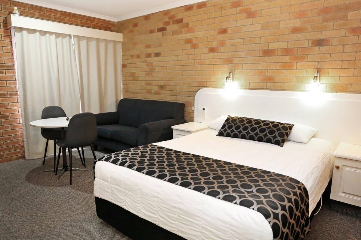 A hotel room with a brick wall, a bed with a patterned bedspread, a desk with a chair, and a window with curtains.